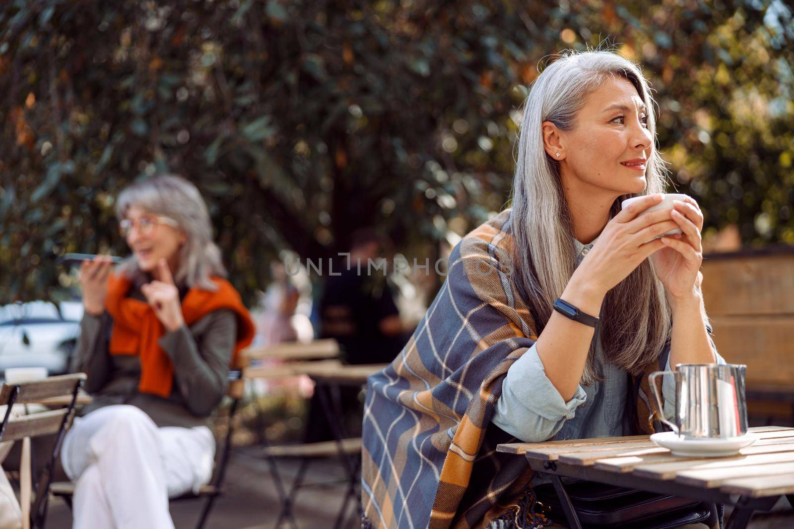 Senior cafe guests, focus on thoughtful hoary haired mature woman holding cup at table on outdoors cafe terrace on autumn day