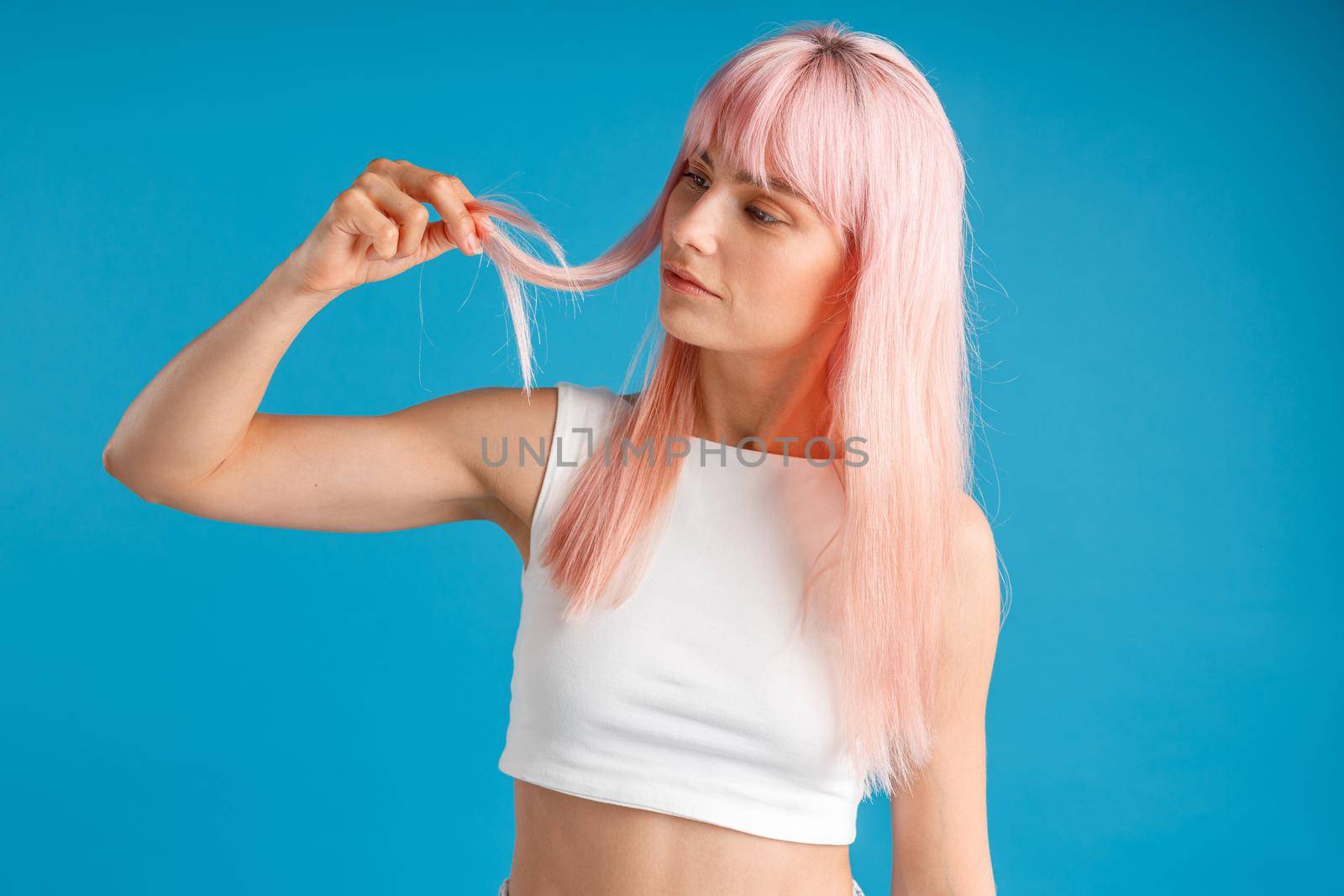 Curious woman touching and looking at her smooth natural long pink dyed hair while posing isolated over blue studio background. Beauty, hair care concept