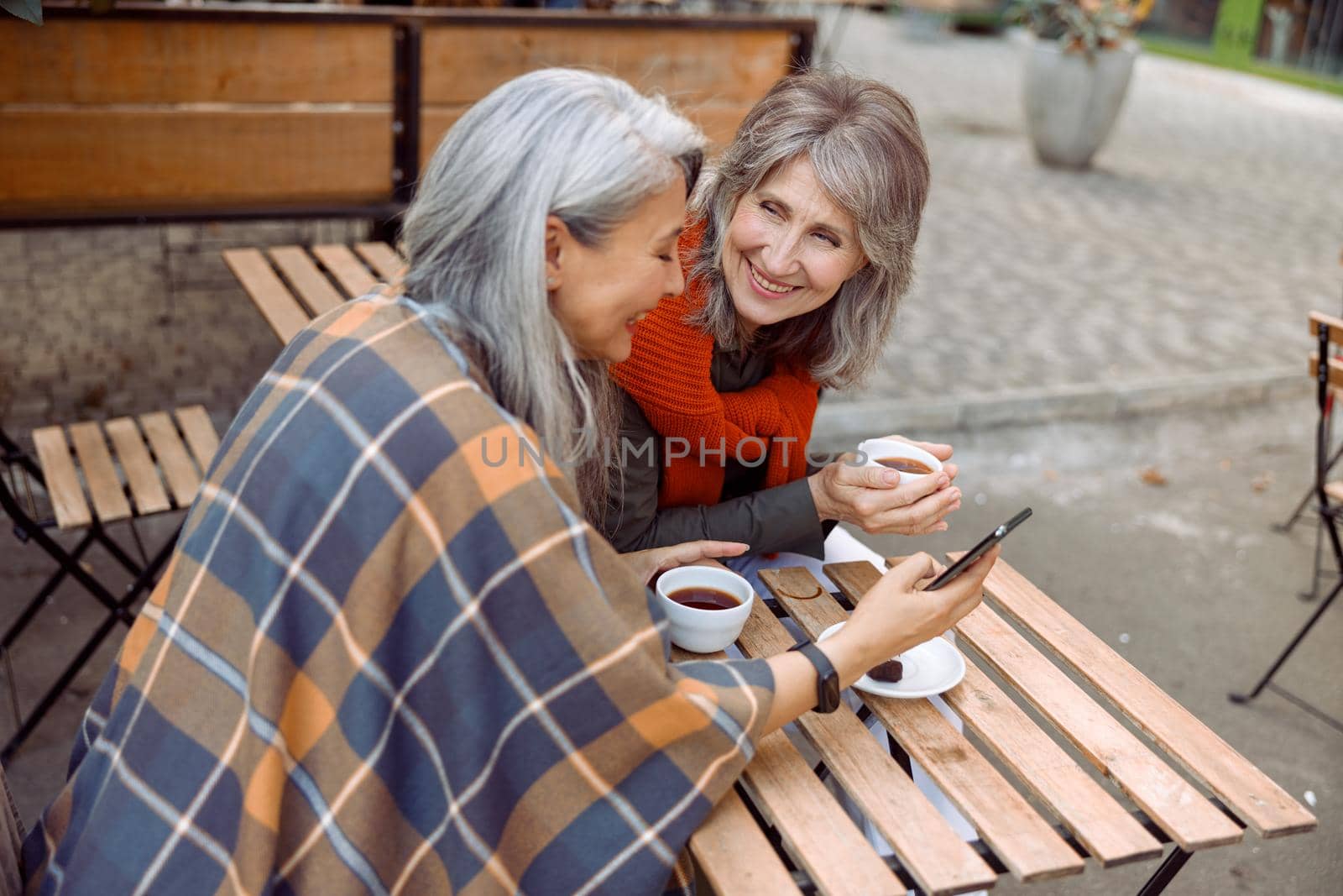 Long haired mature Asian woman shows mobile phone to best friend with cup of coffee at small table in street cafe on autumn day