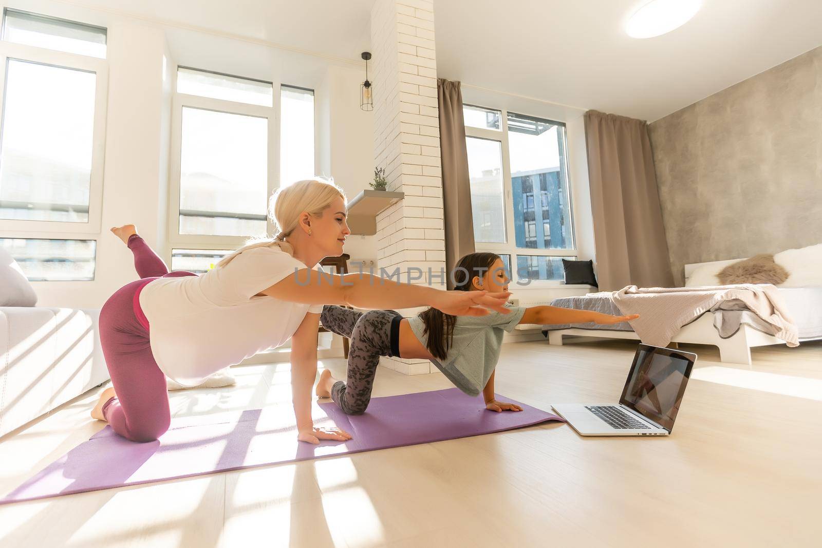 Mother and daughter practicing online yoga classes at home during the quarantine during the coronavirus pandemic. Family doing sports together online from home during isolation together at home by Andelov13