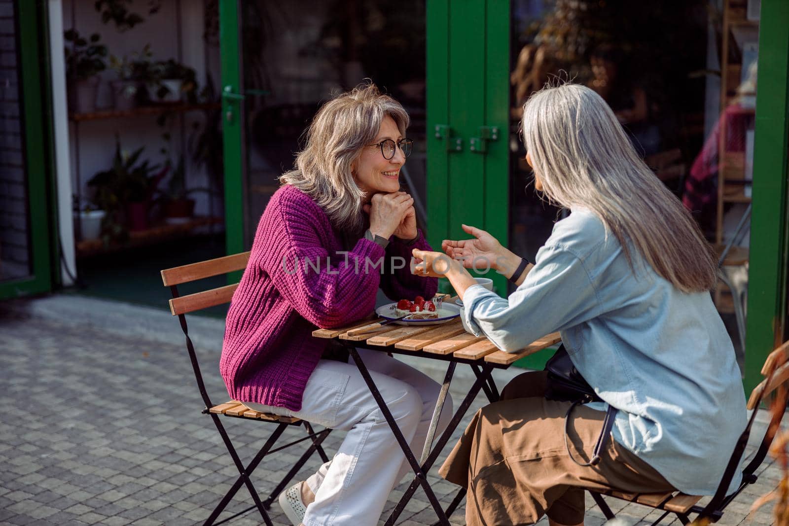 Silver haired woman tells story to mature friend sitting at small table with dessert on outdoors cafe terrace on autumn day