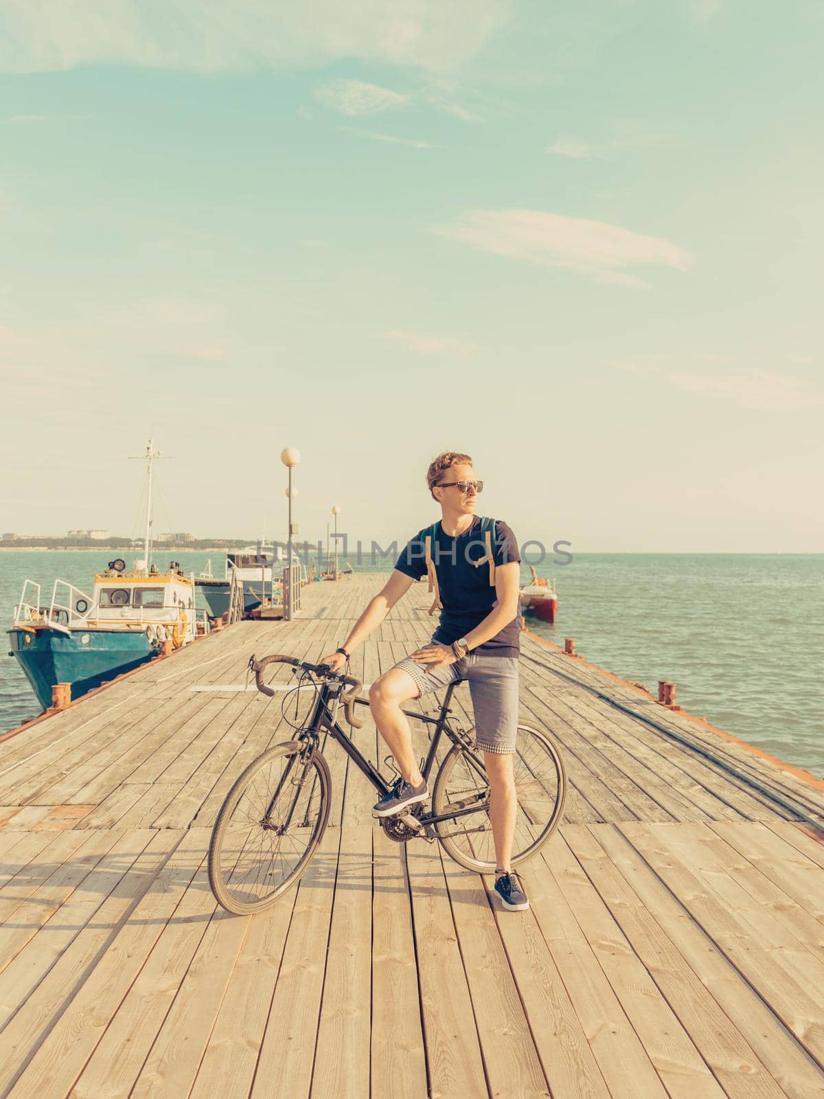 Handsome guy standing with urban bicycle on wooden pier on background of sea in summer. Toned image.