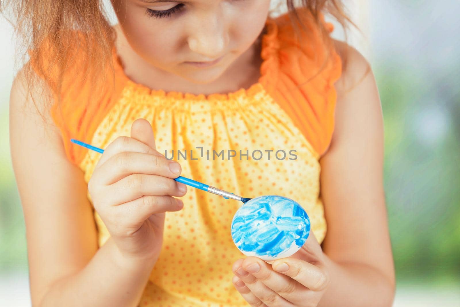 Cute little girl paints egg in blue color for Easter day indoors