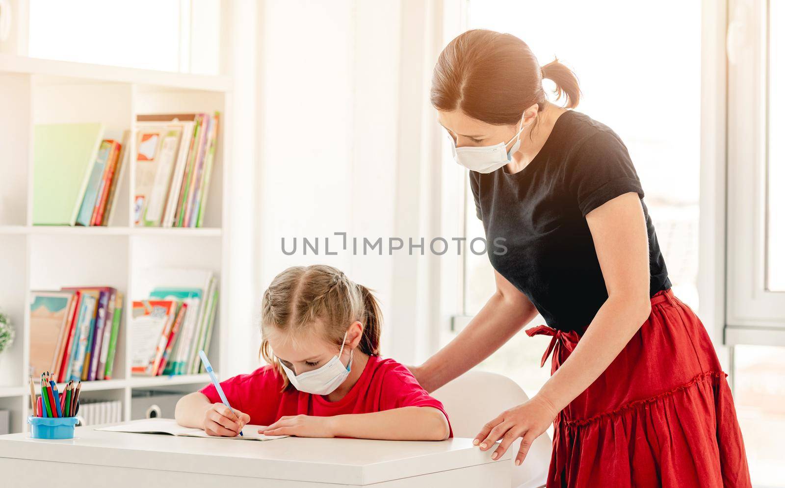 Little girl writing in notebook with teacher's help at school during virus epidemic