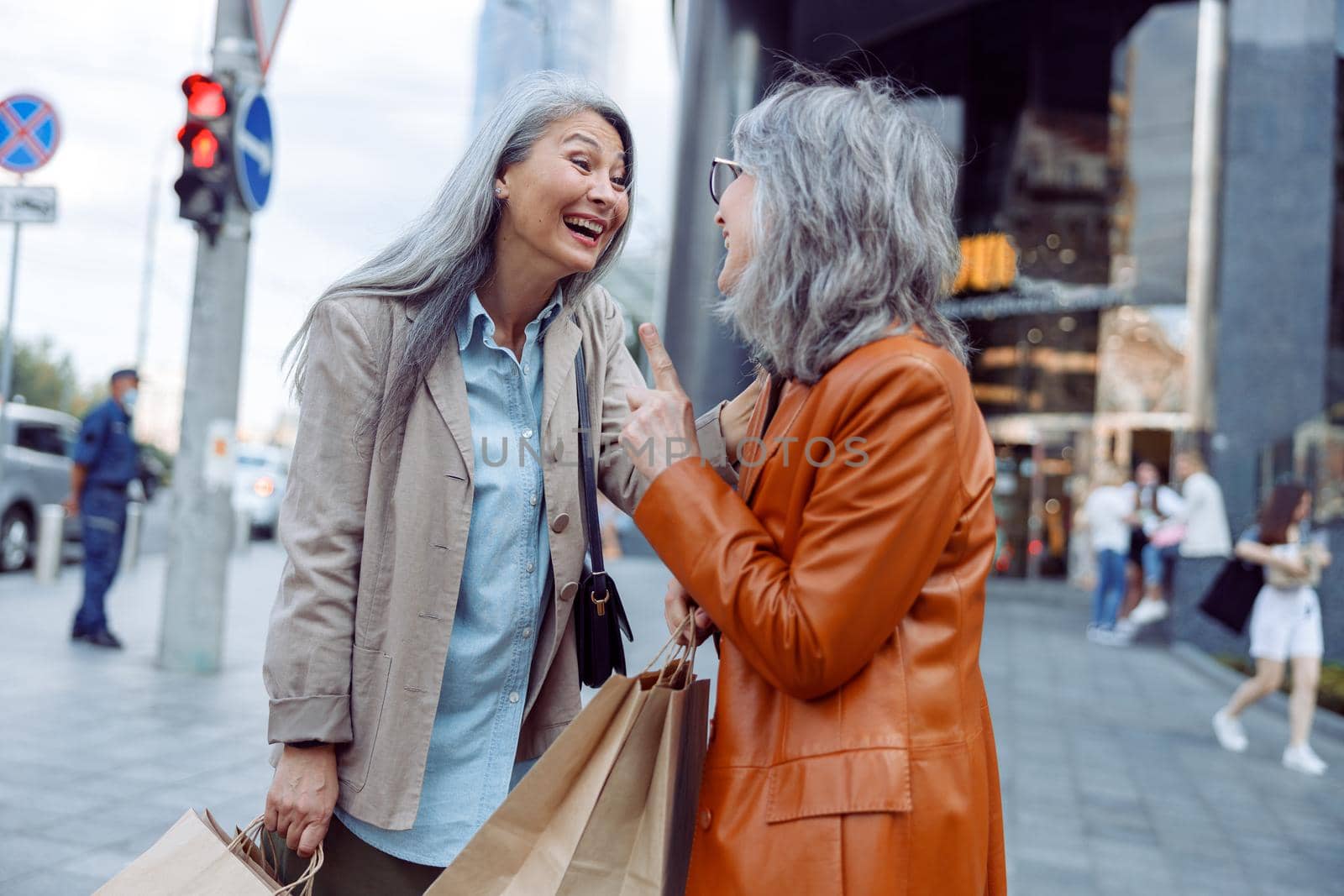 Cheerful mature women in stylish outfit with shopping bags talk on modern city street on autumn day. Friends spend time together