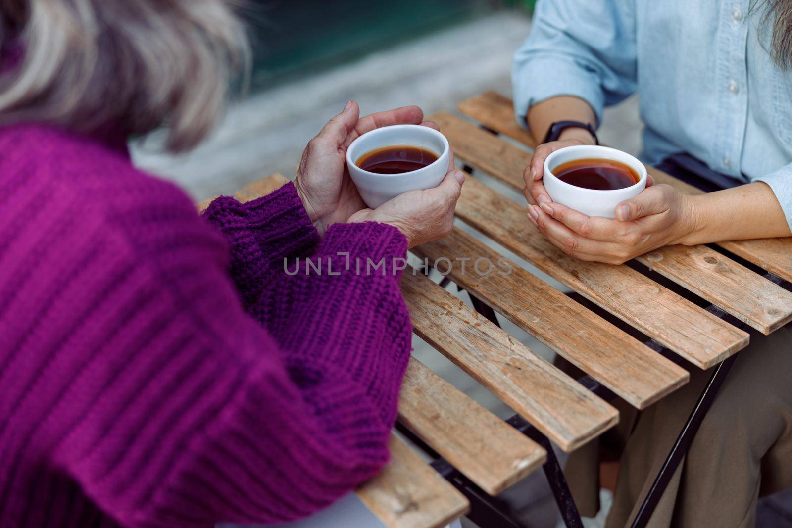 Mature women best friends with cups of fresh coffee sit at wooden table on outdoors cafe terrace on autumn day closeup