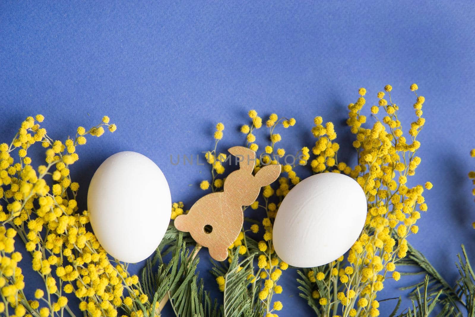 Easter background, white eggs and a wooden hare on a blue background, decorated with Mimosa flowers, flatlay, top view, empty space for text. happy Easter.