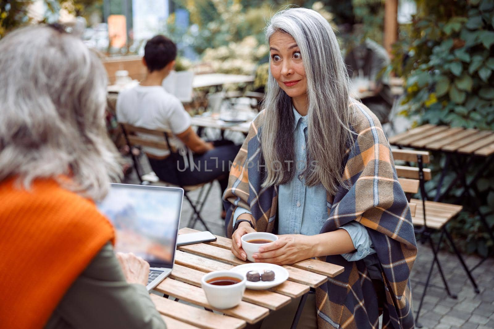 Surprised mature Asian lady looks at friend using laptop while sitting together at small table in street cafe on nice autumn day