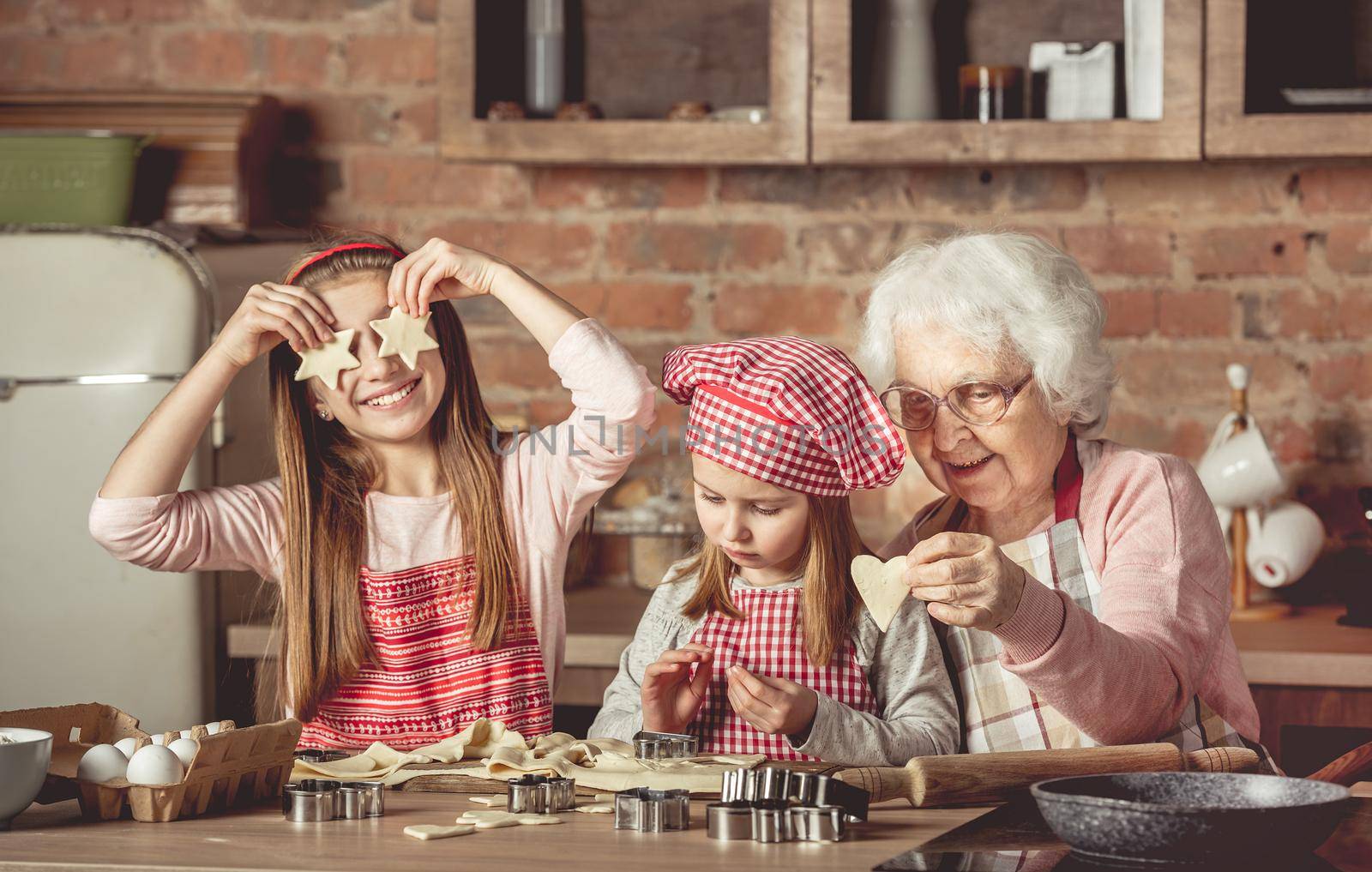 Grandma teaching granddaughters to bake homemade cookies. Little girl holding two star-shaped cookies in front of her eyes and smiling