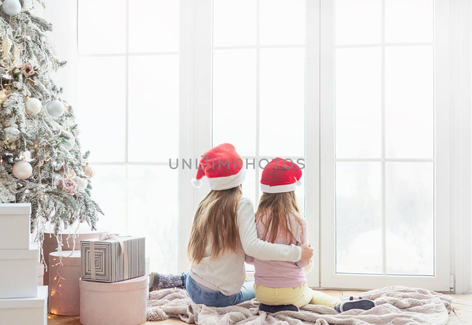 Sisters in santa hats sit hugging near christmas tree