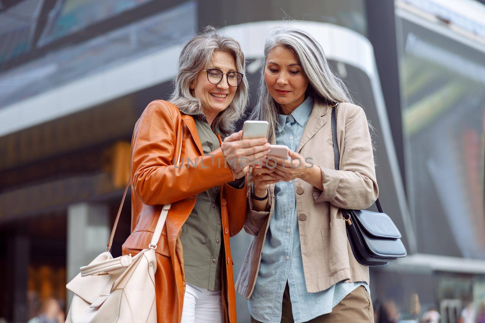 Positive grey haired mature ladies look at modern mobile phones on modern city street on nice autumn day. Old friends meeting