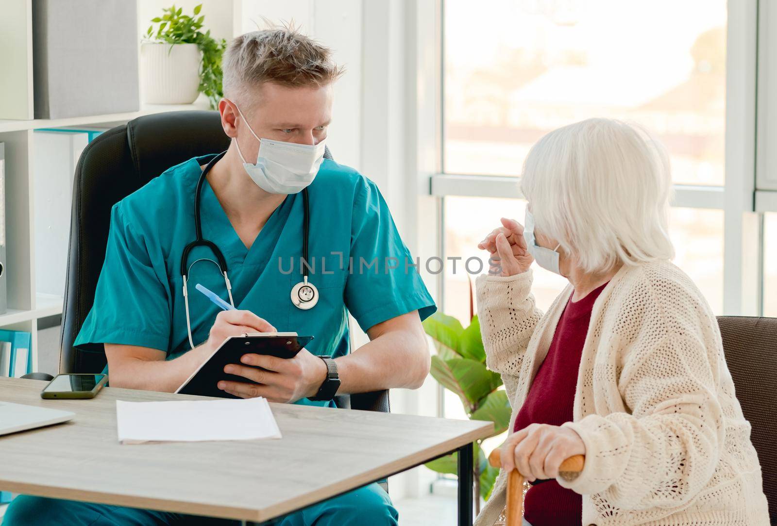 Doctor in protective face mask writing elderly patient complaints during appointment in clinic