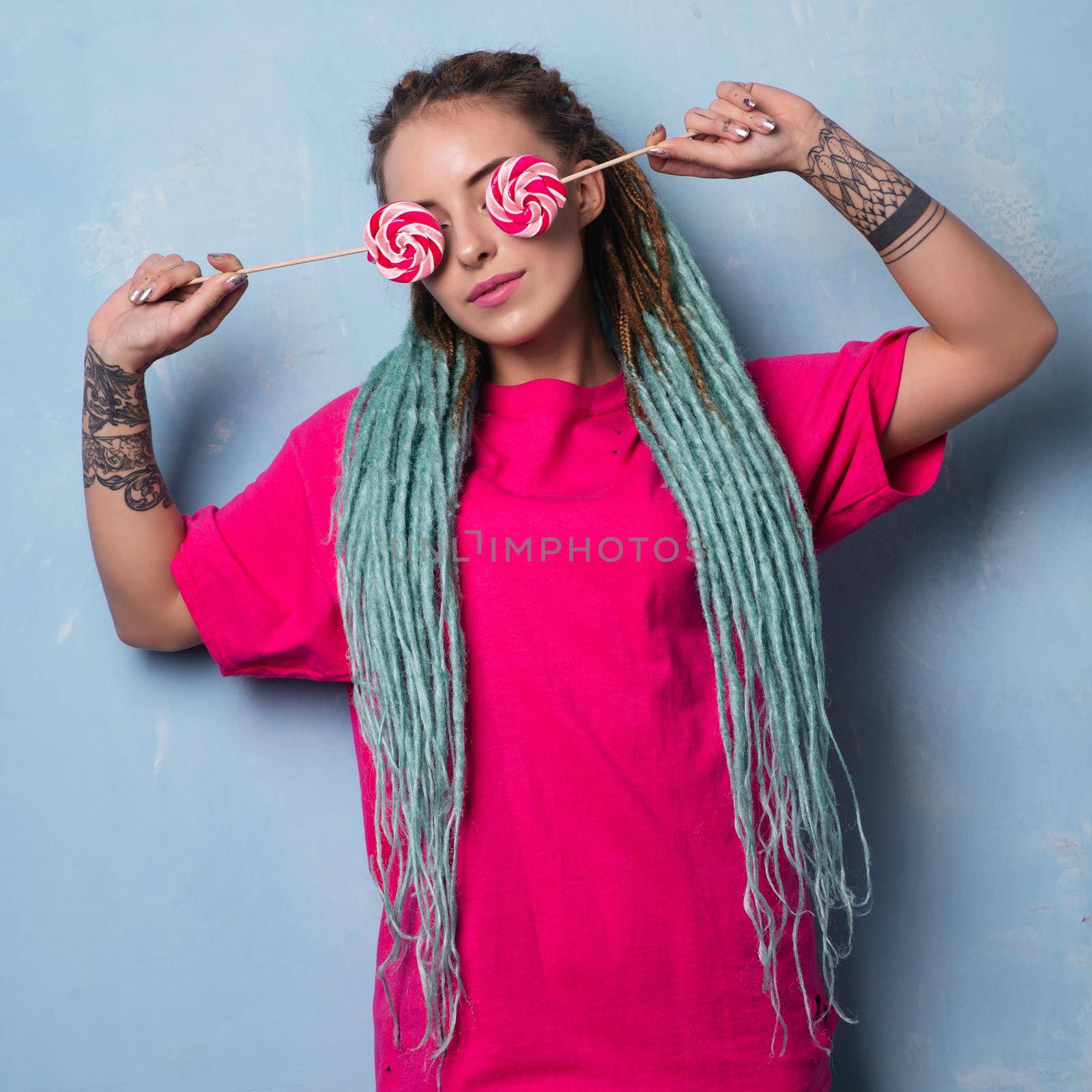 Cute girl in pink t-shirt with dreadlocks and tattoo closes her eyes with lollipops.