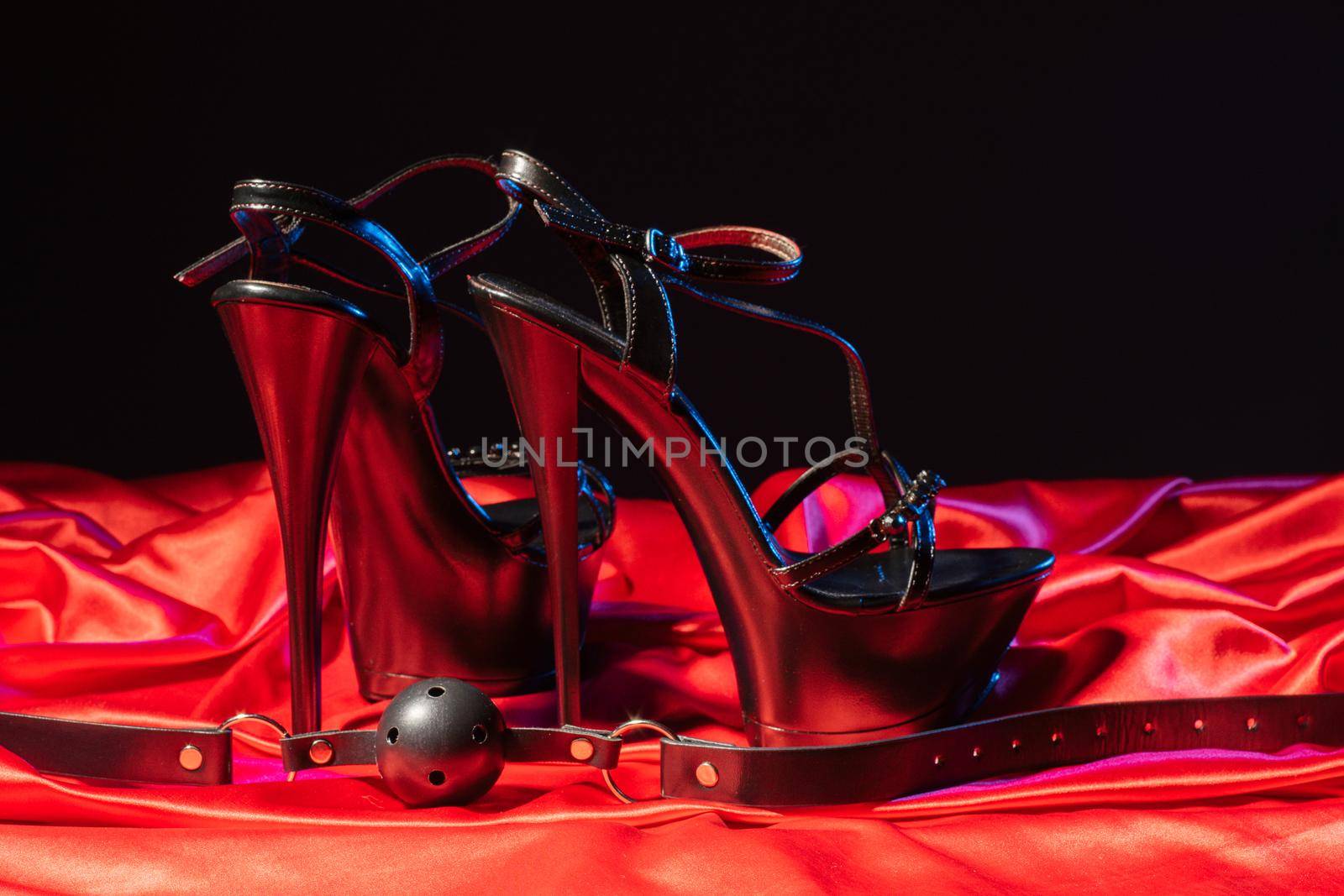 Adult sex games. Kinky lifestyle. Gag ball and a pair of black high-heeled shoes on the red linen. Bdsm outfit - Image