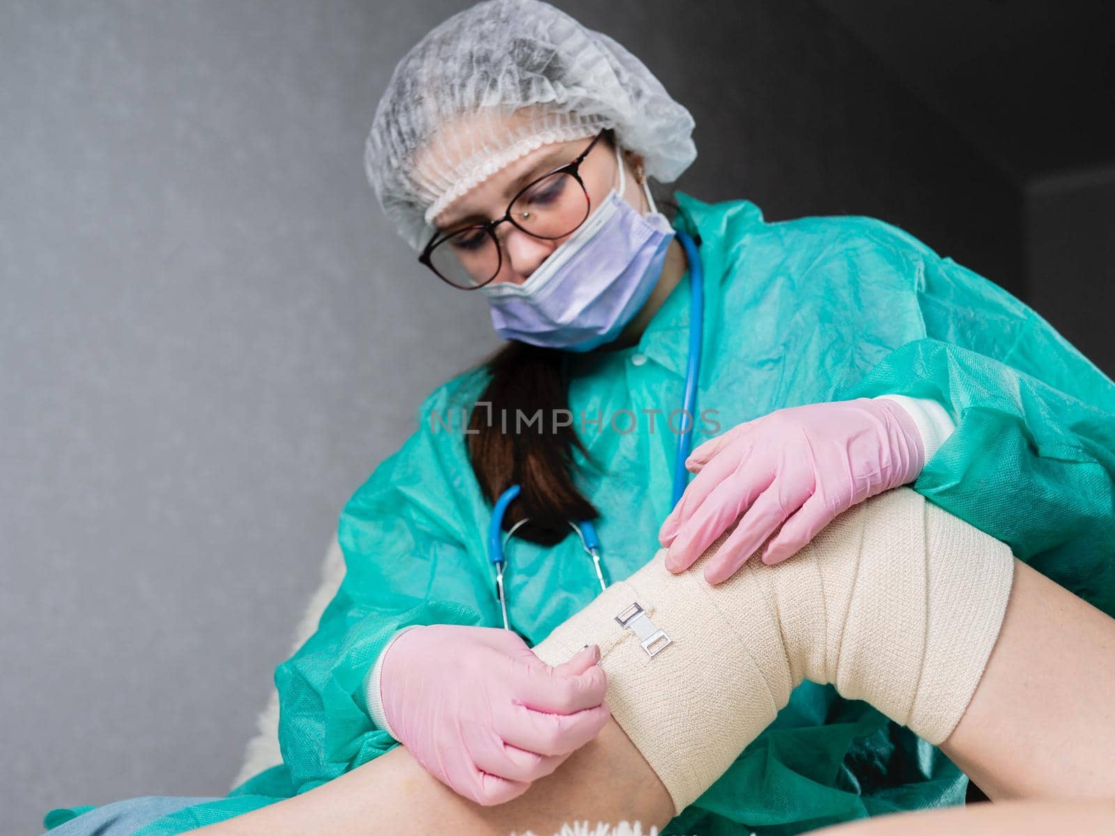 A young nurse wearing medical gloves puts an elastic bandage on the patient's leg and knee. Physiotherapy room. by Utlanov