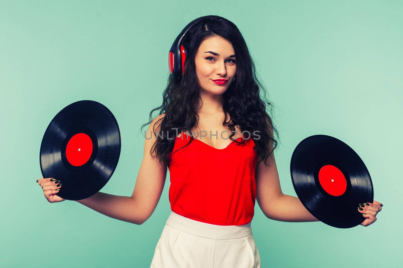Young woman in headphones looking at camera and closing her face with vinyl record on blue background. - Image Toned