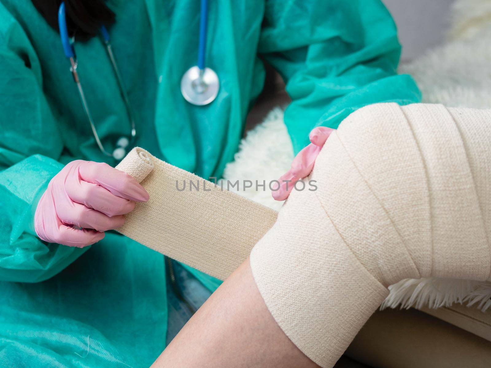 A young nurse wearing medical gloves puts an elastic bandage on the patient's leg and knee. Physiotherapy room.