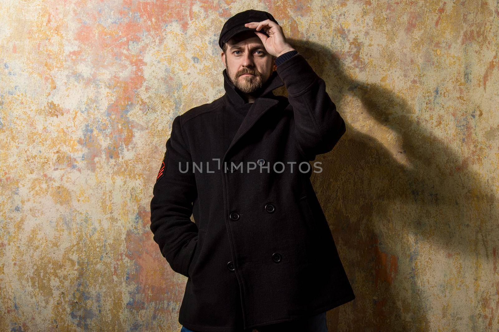 attractive young bearded man in a pea coat and cap is standing at the wall