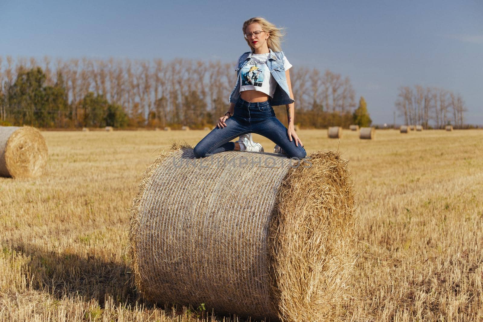 Beautiful girl villager posing in jeans on a bale of hay in a field by zartarn