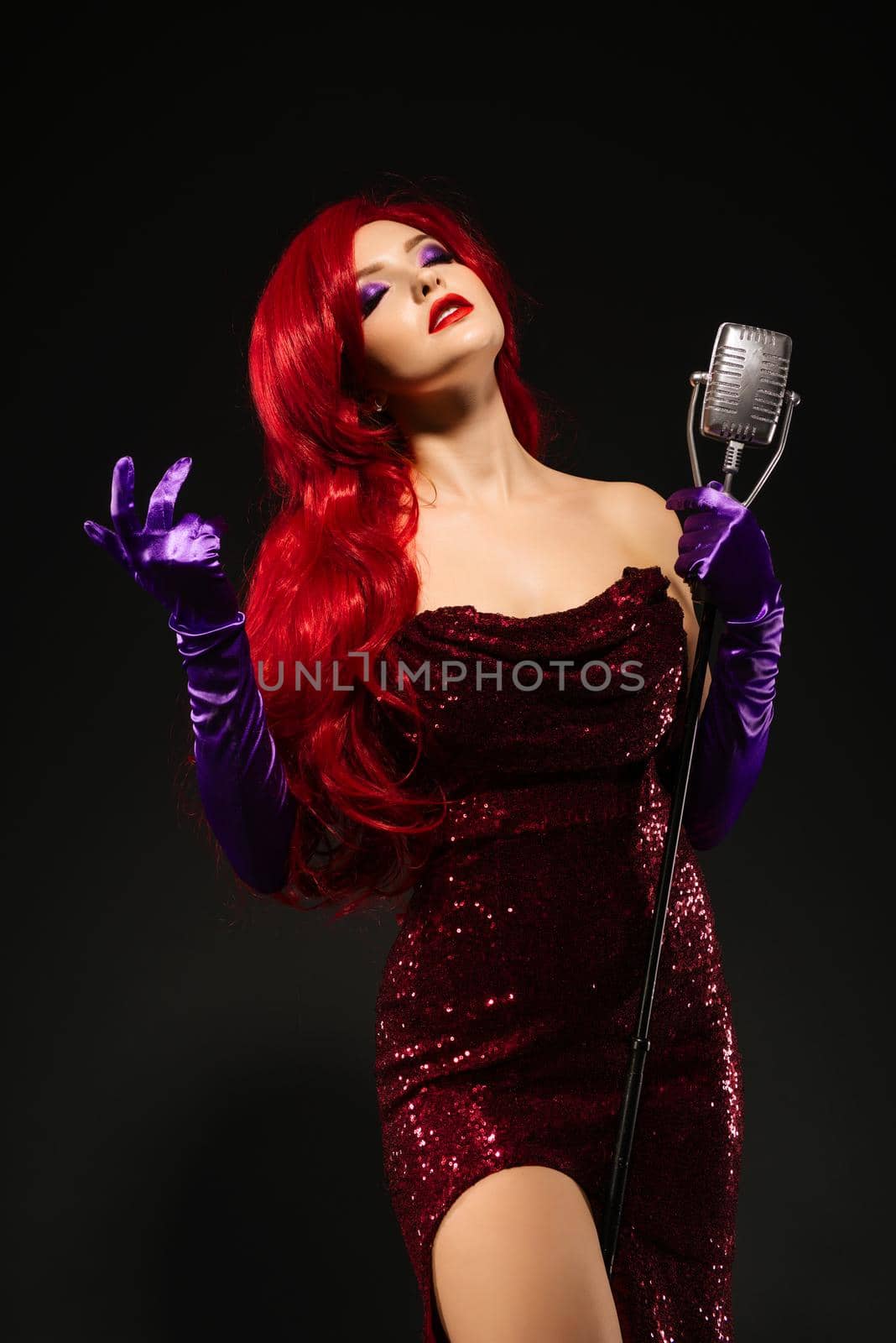 Young romantic redhead woman with very long hair in red dress with microphone on the stand sings with his eyes closed on a black background. by zartarn