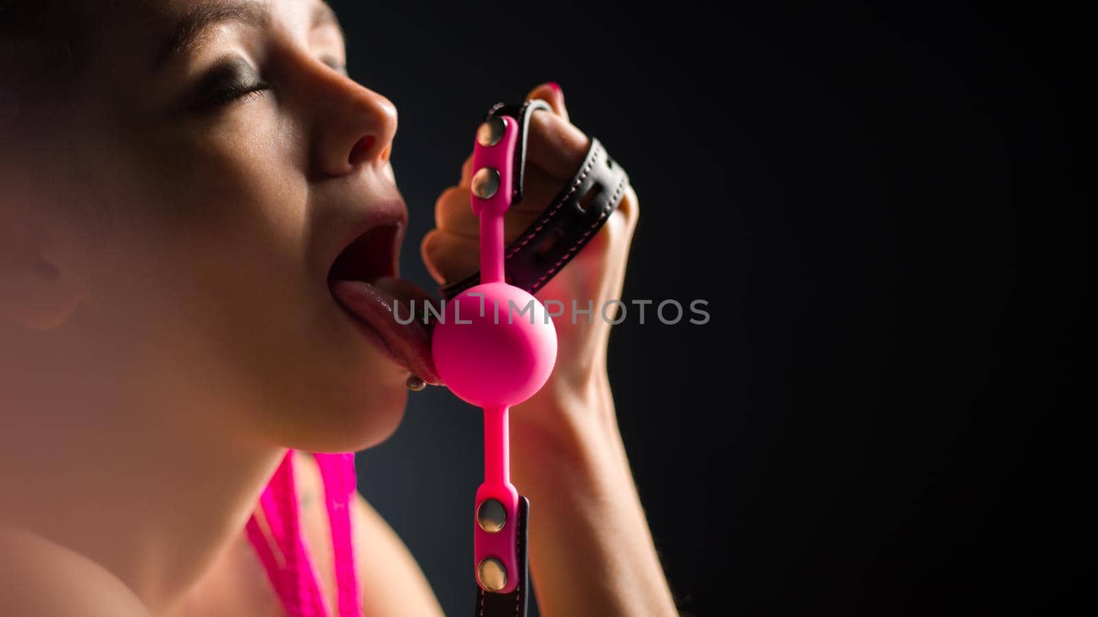 BDSM outfit for adult sex games. A young woman licks pink gag ball - image
