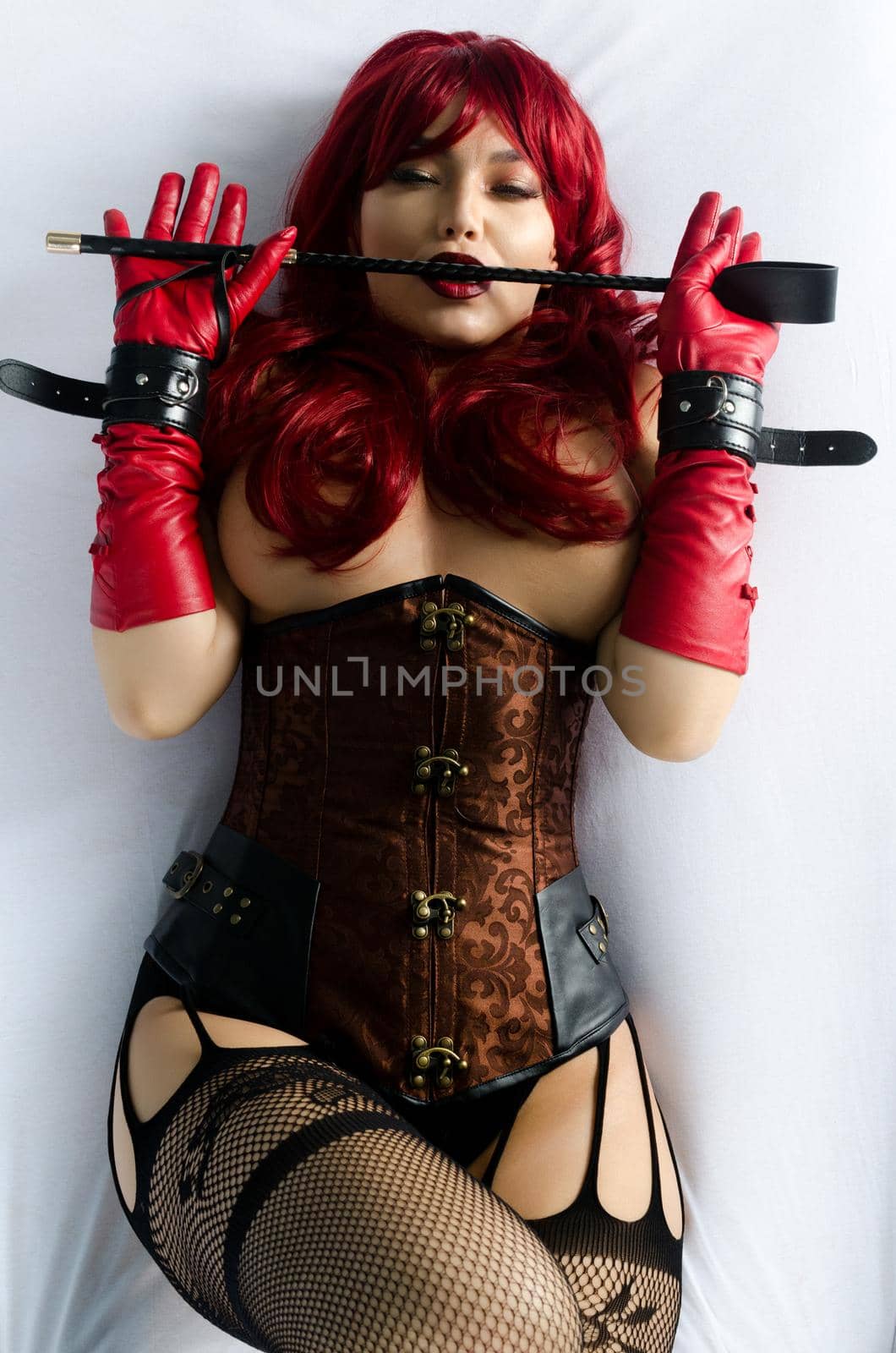 Red-haired woman in a red wig, corset and leather gloves posing lying on a bed holding a stick in the hands of a whip by zartarn