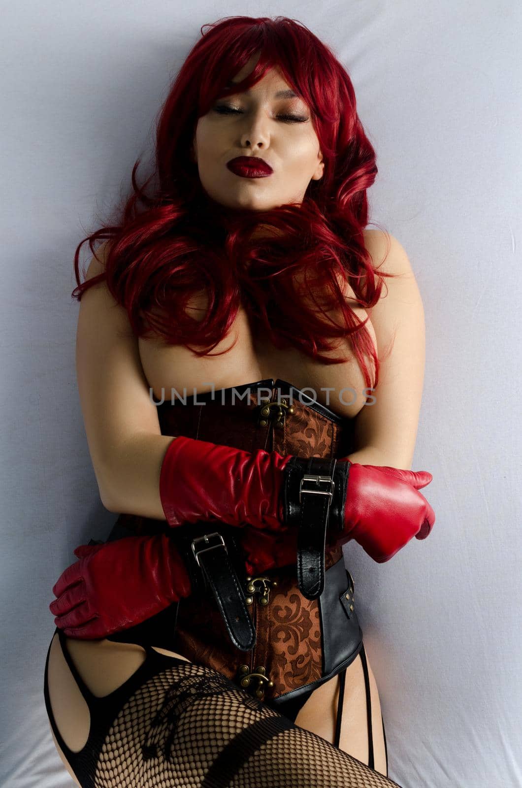 Red-haired woman in a red wig, corset and leather gloves posing seductively lying on the bed. Adult sex game