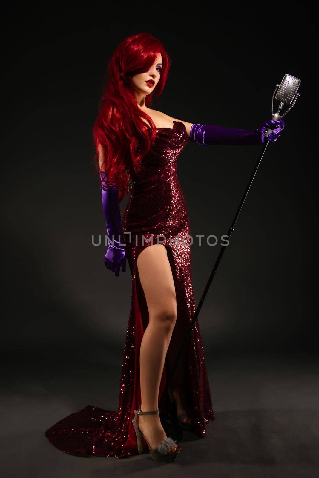 Full-length portrait of young romantic redhead woman with very long hair in red dress with microphone on the stand on a black background. Sexy gown on a beautiful slim figure. Long pink gloves