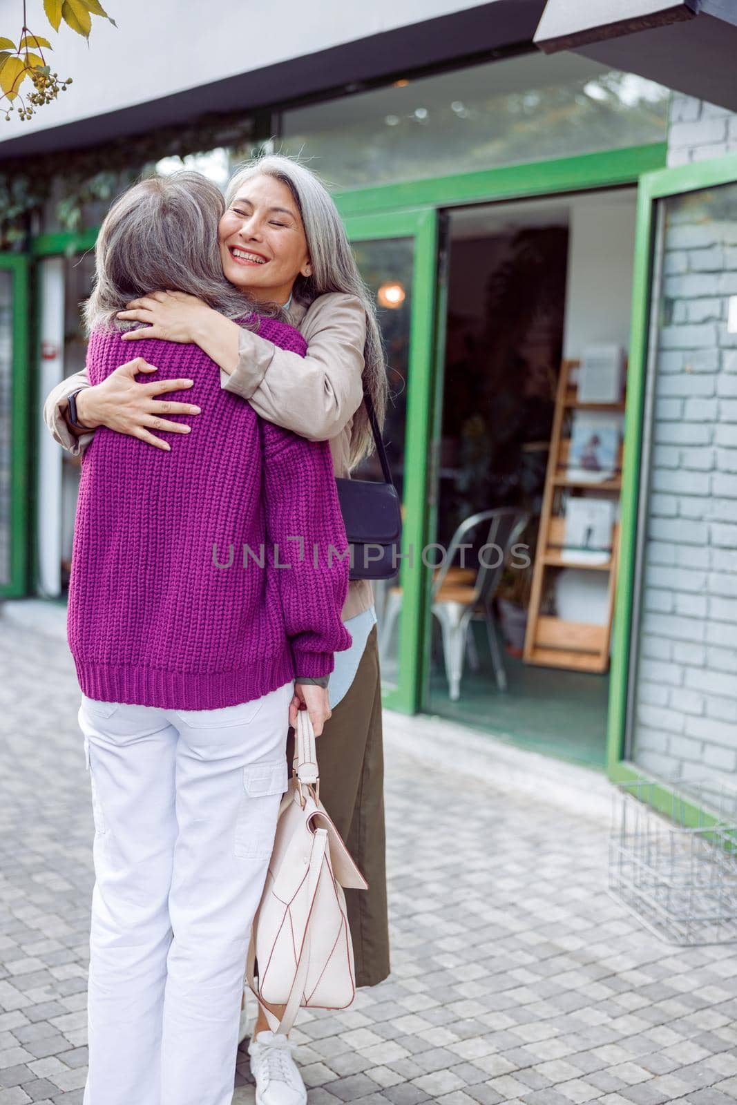 Happy senior Asian lady hugs best friend wearing purple knitted jacket meeting on modern city street. Long-time friendship relationship