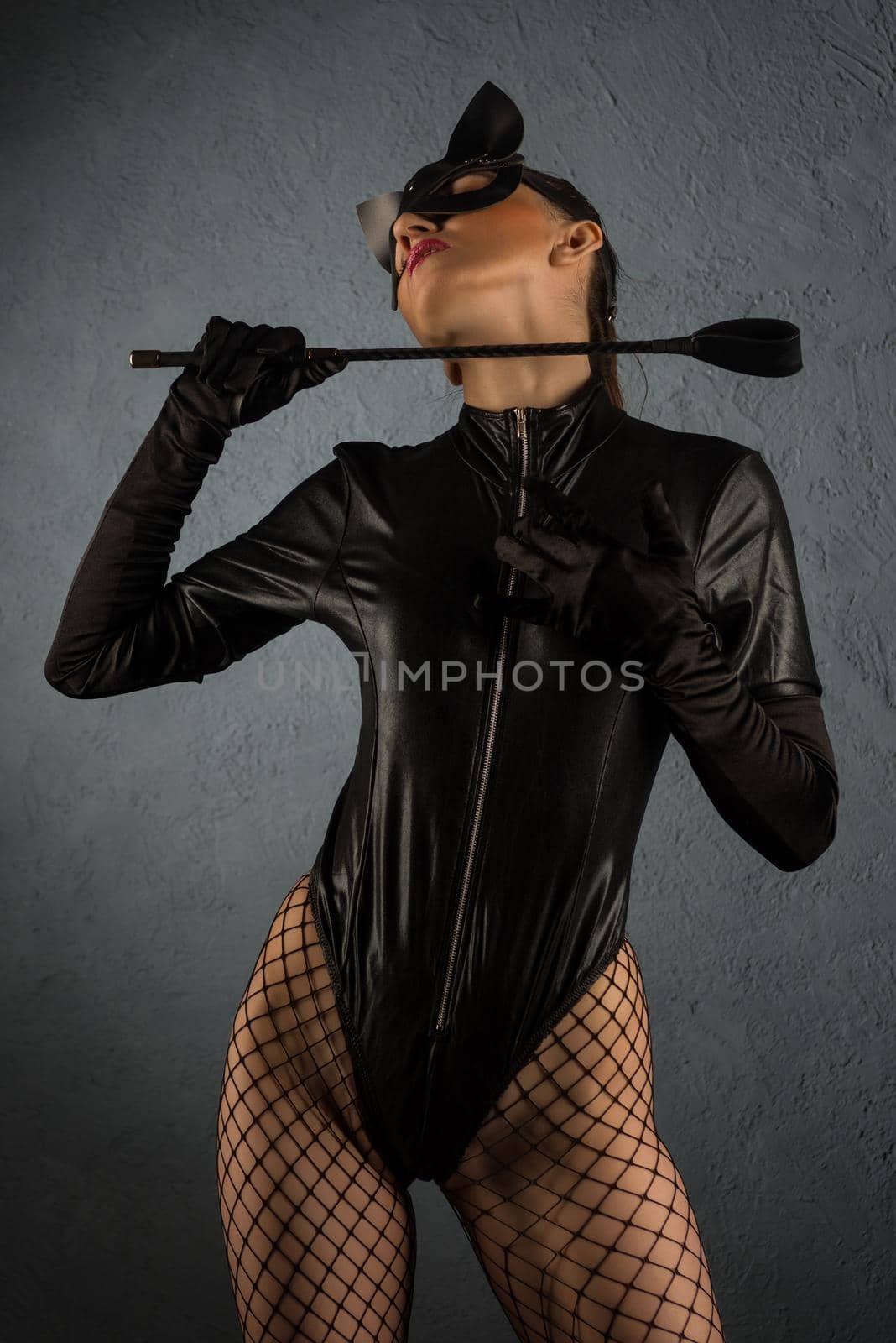 Adult sex games. Beautiful dominant brunette vamp mistress girl in latex body, gloves and bdsm black leather fetish cat mask posing with riding crop. - image