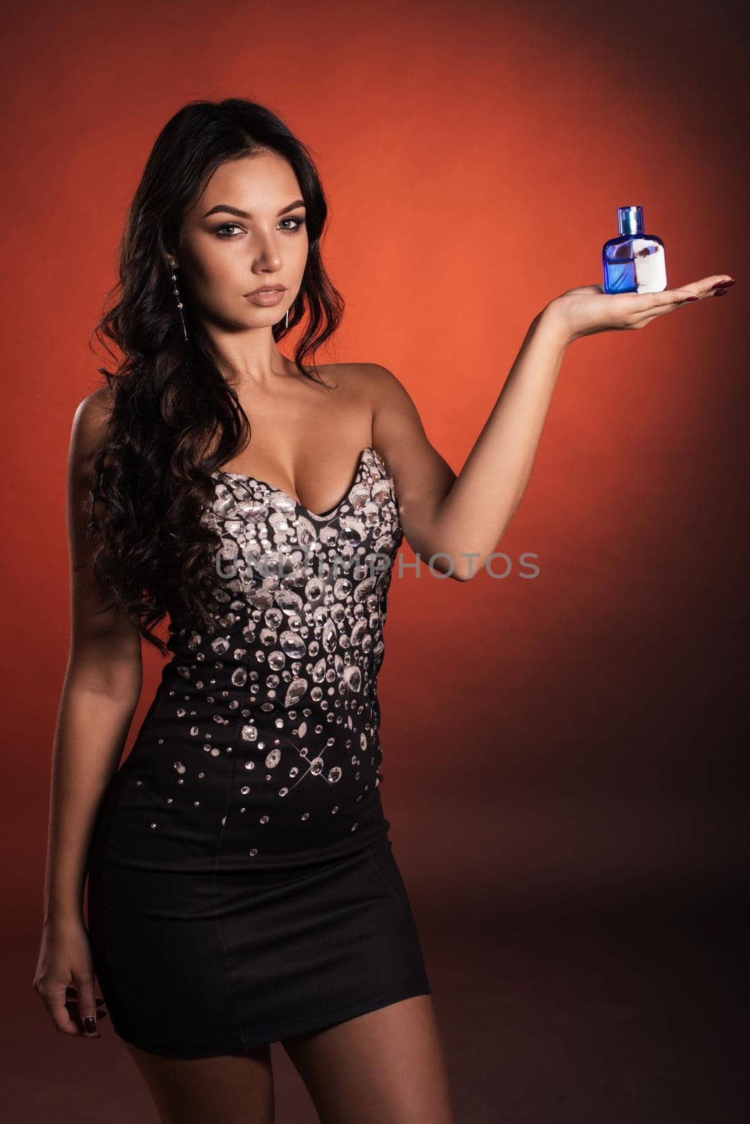 Beautiful luxurious young woman in a dress with rhinestones posing with perfume in a bottle of blue on red background