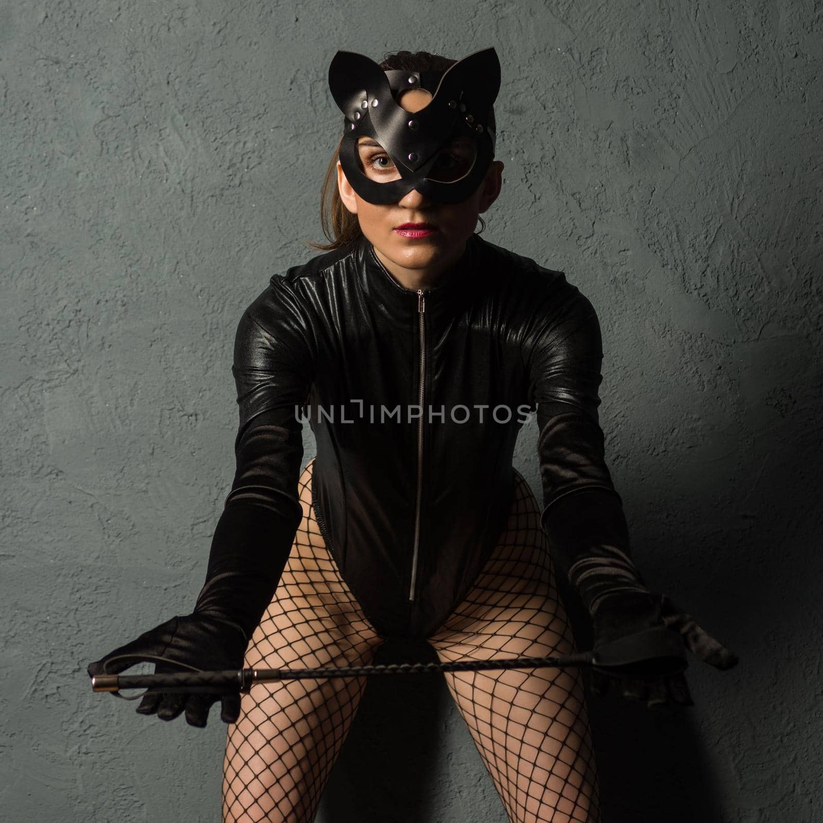 Adult sex games. Beautiful dominant brunette vamp mistress girl in latex body, gloves and bdsm black leather fetish cat mask posing with riding crop. - image