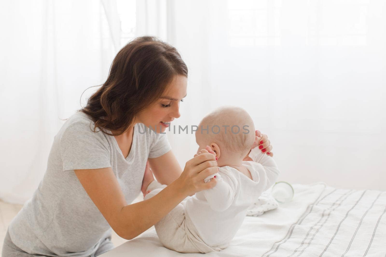 Young mother helping her infant child to sit on bed.