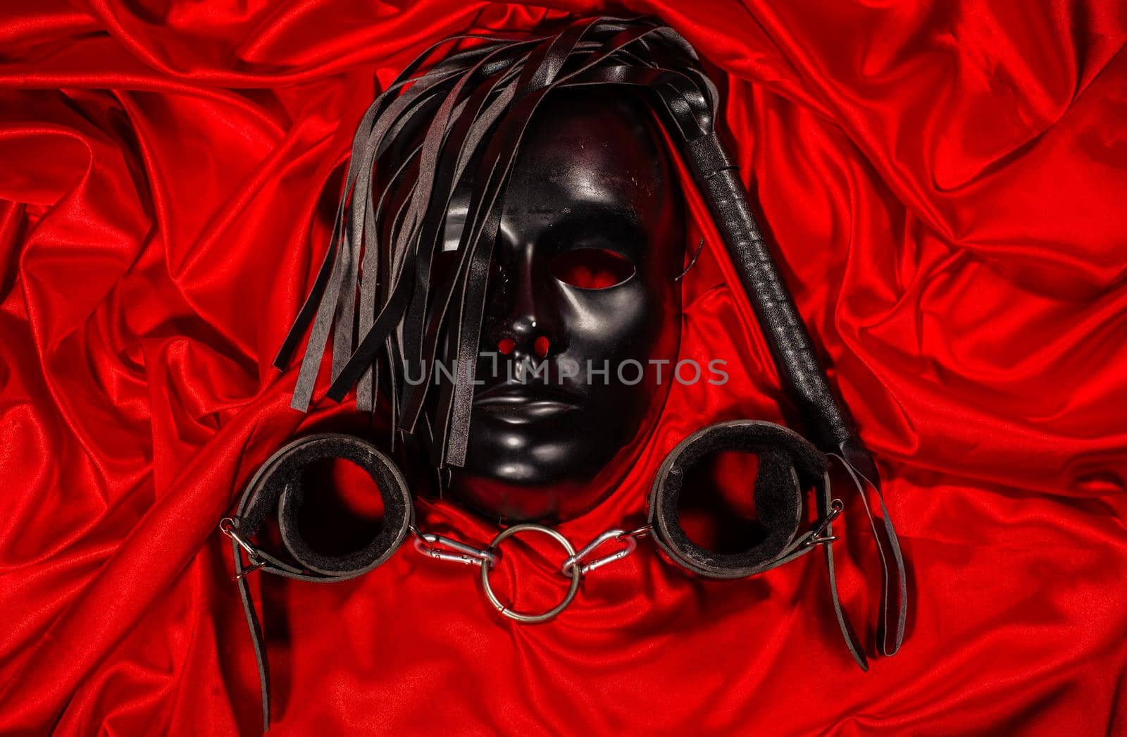 Bondage, kinky adult sex games, kink and BDSM lifestyle concept with a mask, pair of leather handcuffs, flogger, ball gag and a coller with a leash attached on red silk with copy space