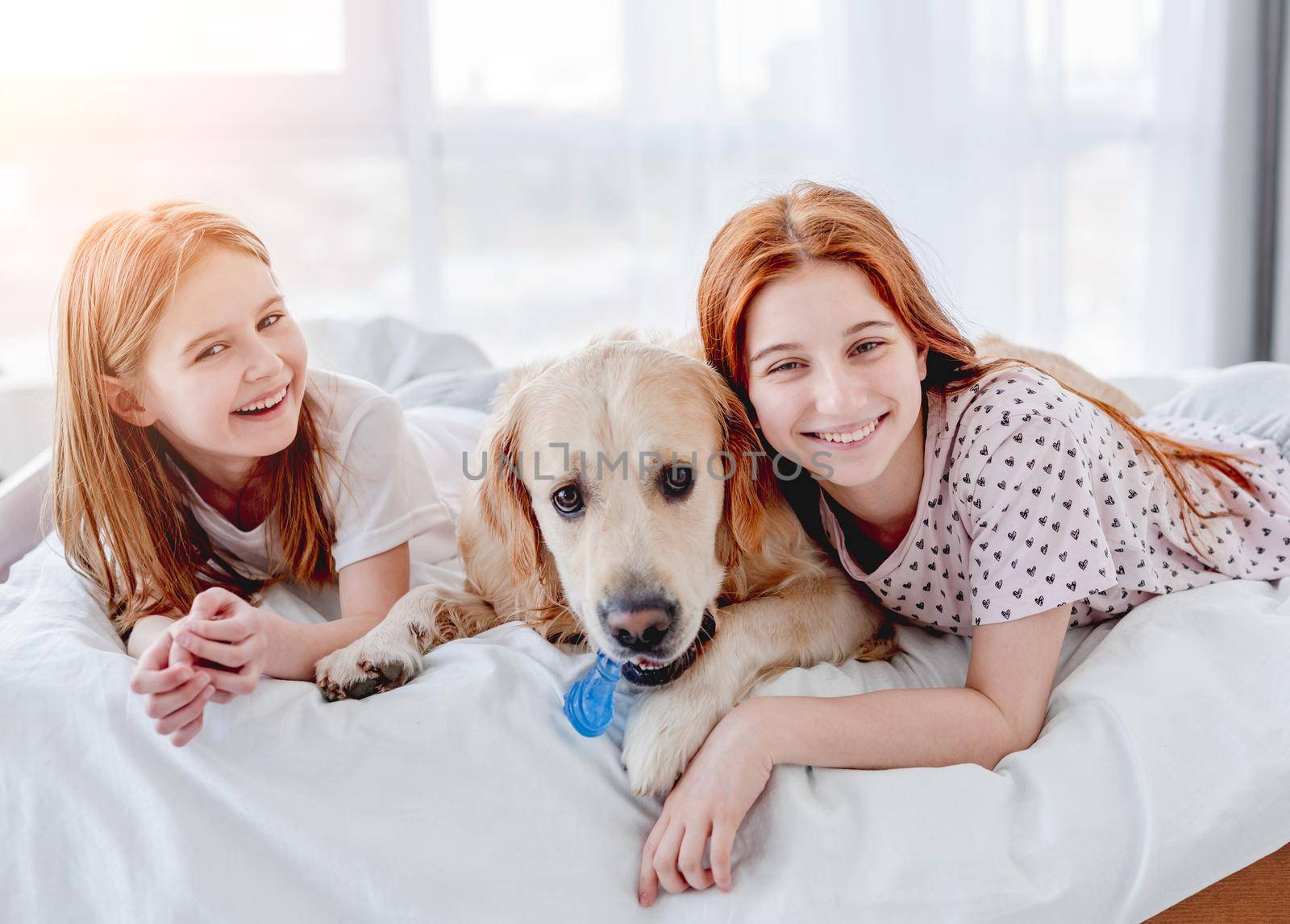 Two smiling sisters lying in the bed with golden retriever dog in the morning time. Girls with pet staying at home. Beautiful portrait of friendship between human and animal doggy