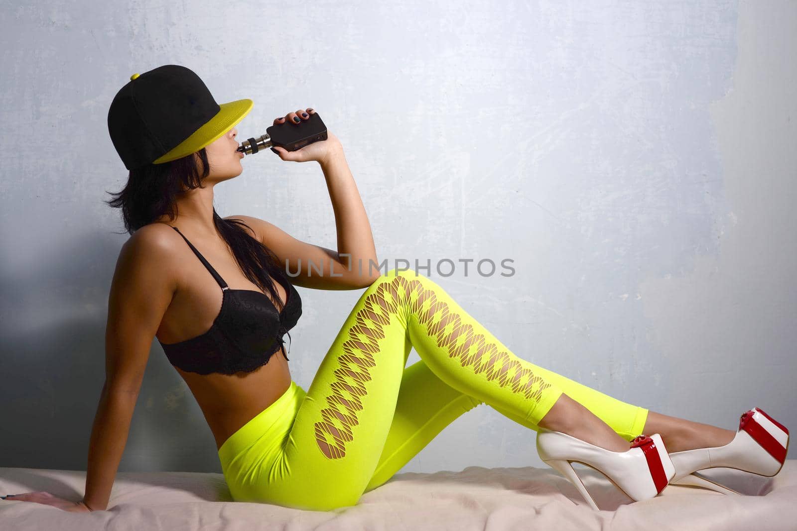 Slim girl in black bra leggings light green high-heeled shoes and a baseball cap smokes electronic cigarette sitting on a red couch