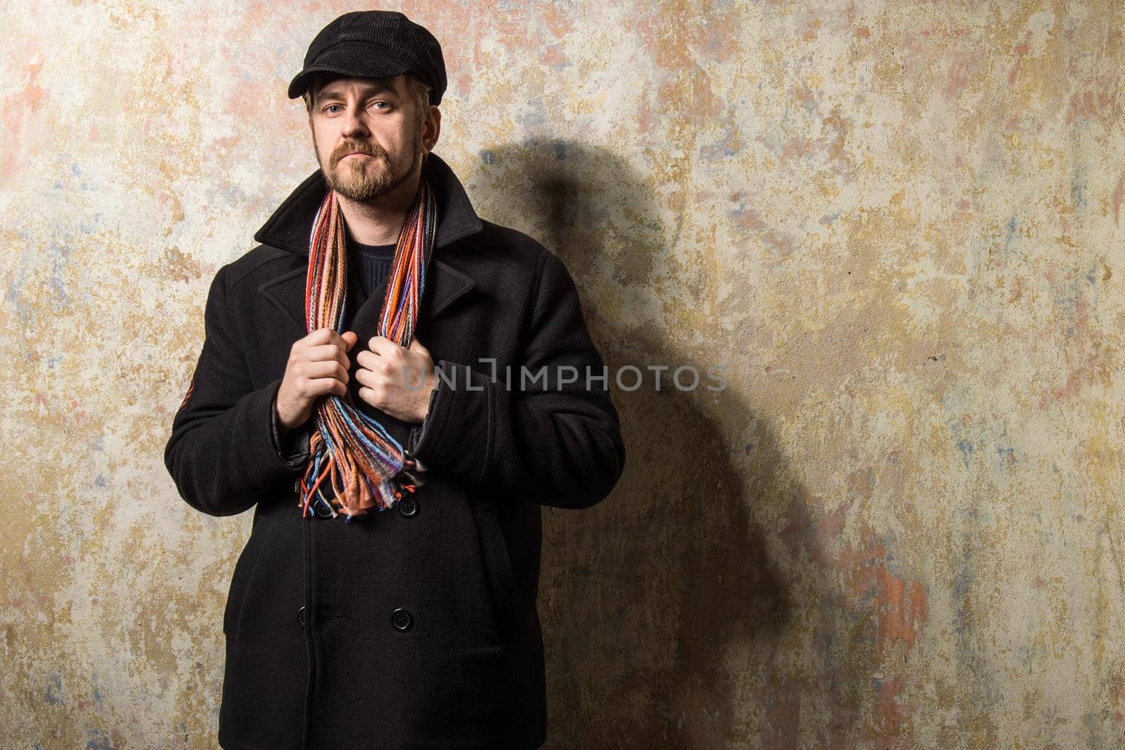 Serious bearded man wearing a cap and autumn coat holding hands to scarf hanging on the neck
