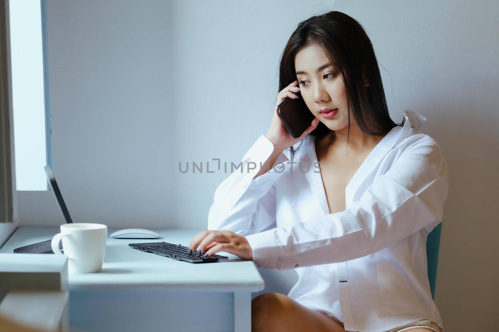 The new normal. A businesswoman is using her phone and computer to work for a company. over the Internet on your desk at home. by Manastrong
