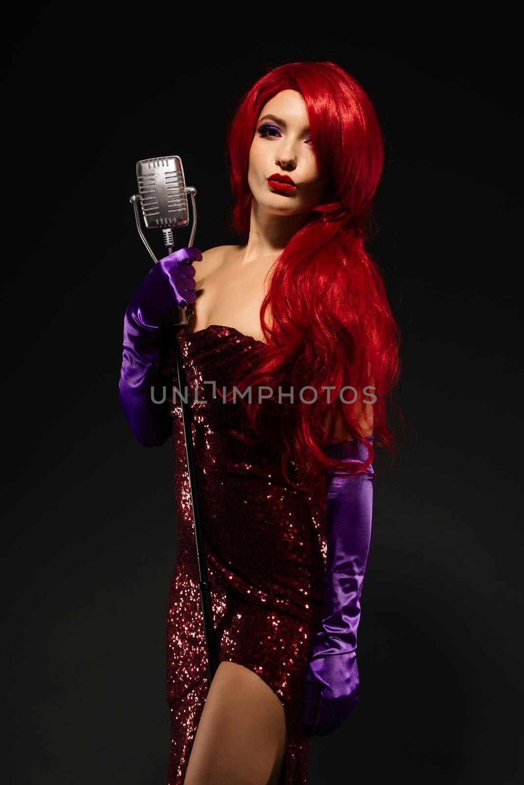 Young romantic redhead woman with very long hair in red dress with microphone on the stand licked lips in a kiss on a black background. by zartarn