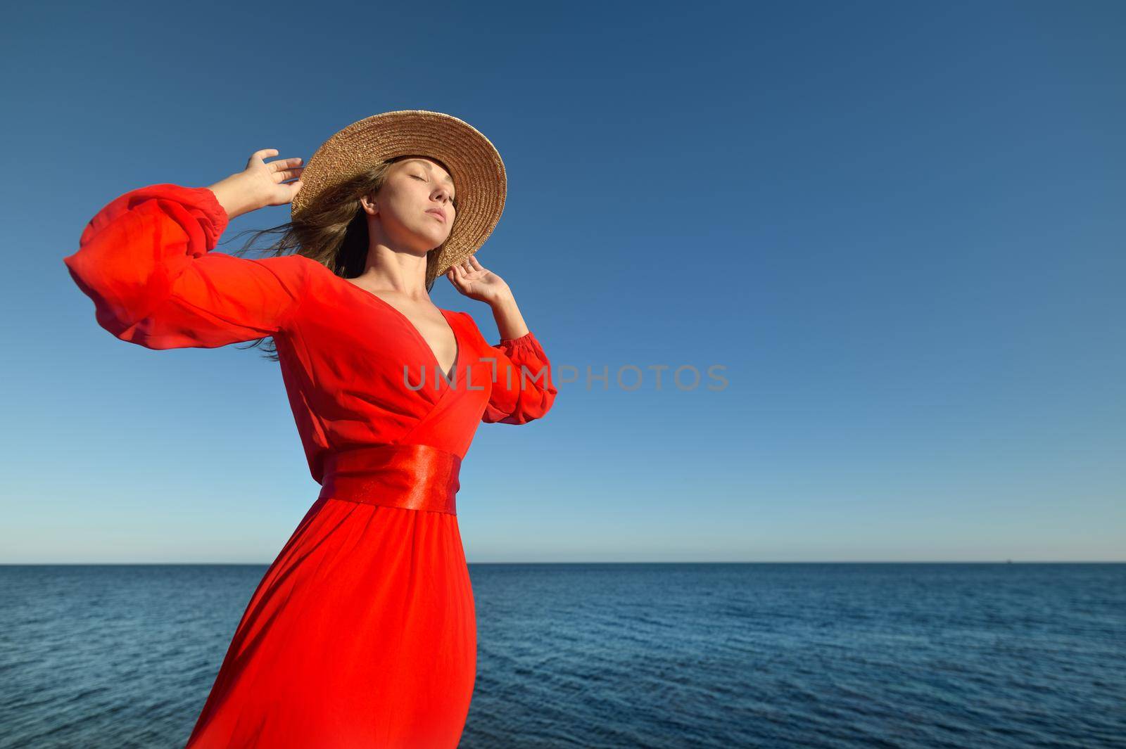 Lovely romantic caucasian young woman in red dress and straw hat against the background of the sea horizon in windy weather.