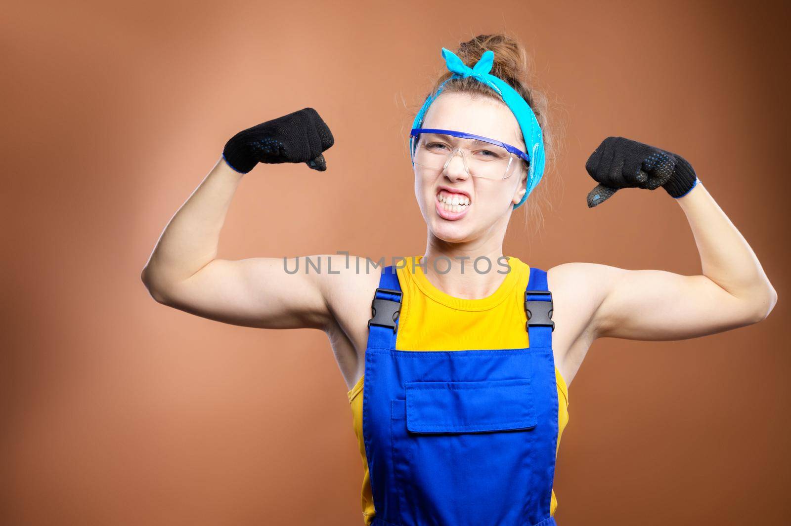 Young beautiful caucasian woman repairman worker with blue eyes in uniform showing arm muscles, proudly smiling and making a strong grimace. The concept of the rapid implementation of industrial projects. Literate contractors and wise construction superintendents