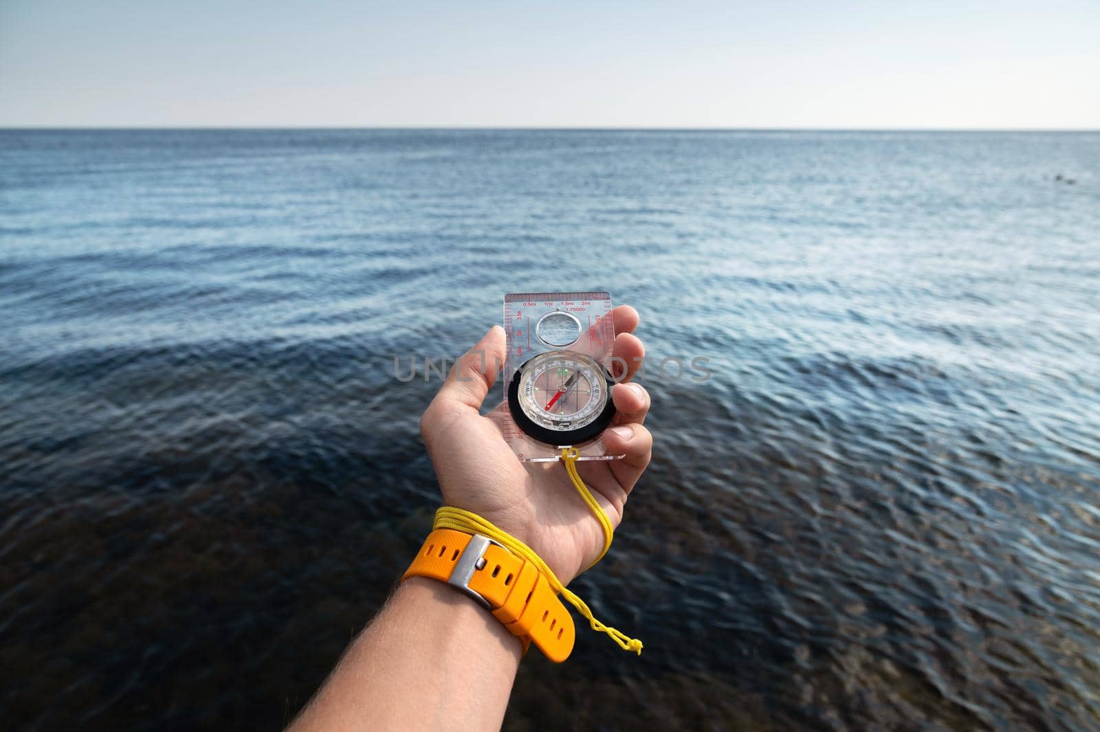 A man's hand with a wristwatch bracelet holds a magnetic compass against the background of the sea.