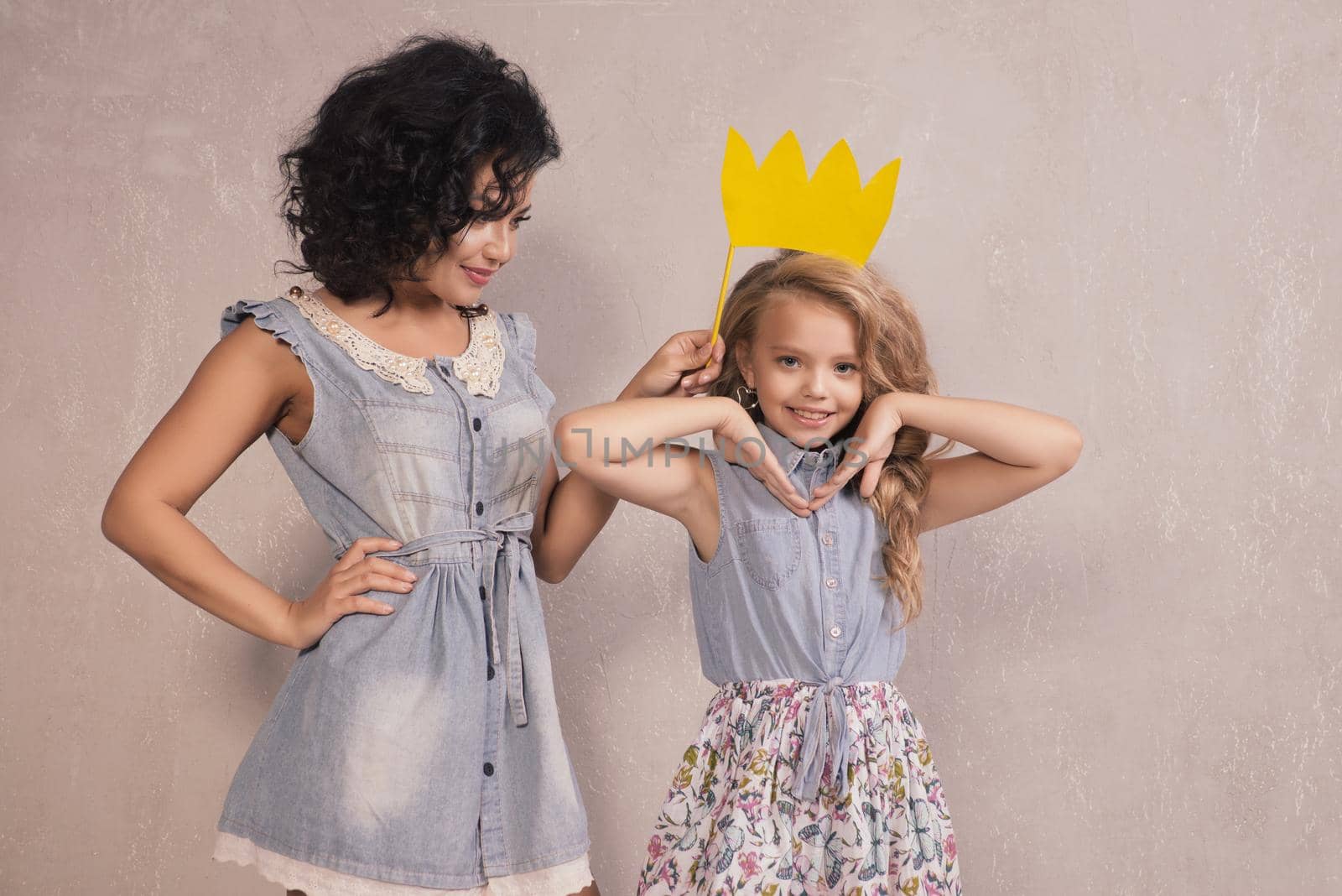 Funny Mother and her daughter girl with yellow paper crown on stick near gray wall