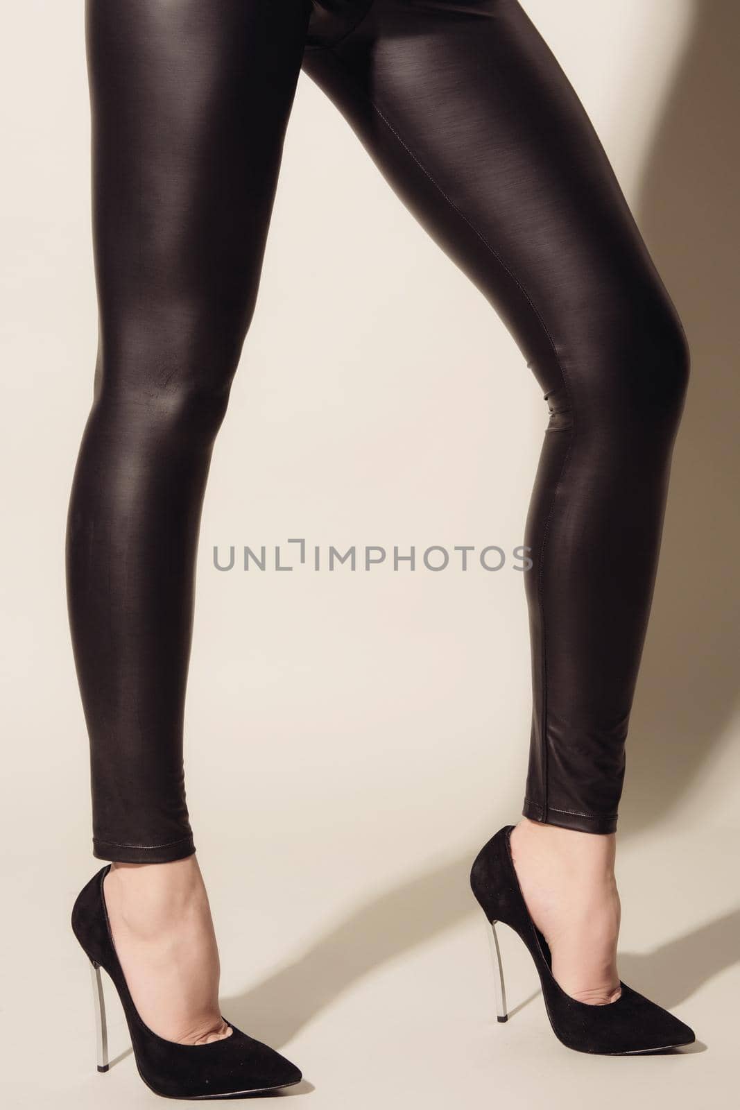 Women's legs in black tight-fitting leather trousers and high-heeled shoes standing on grey background