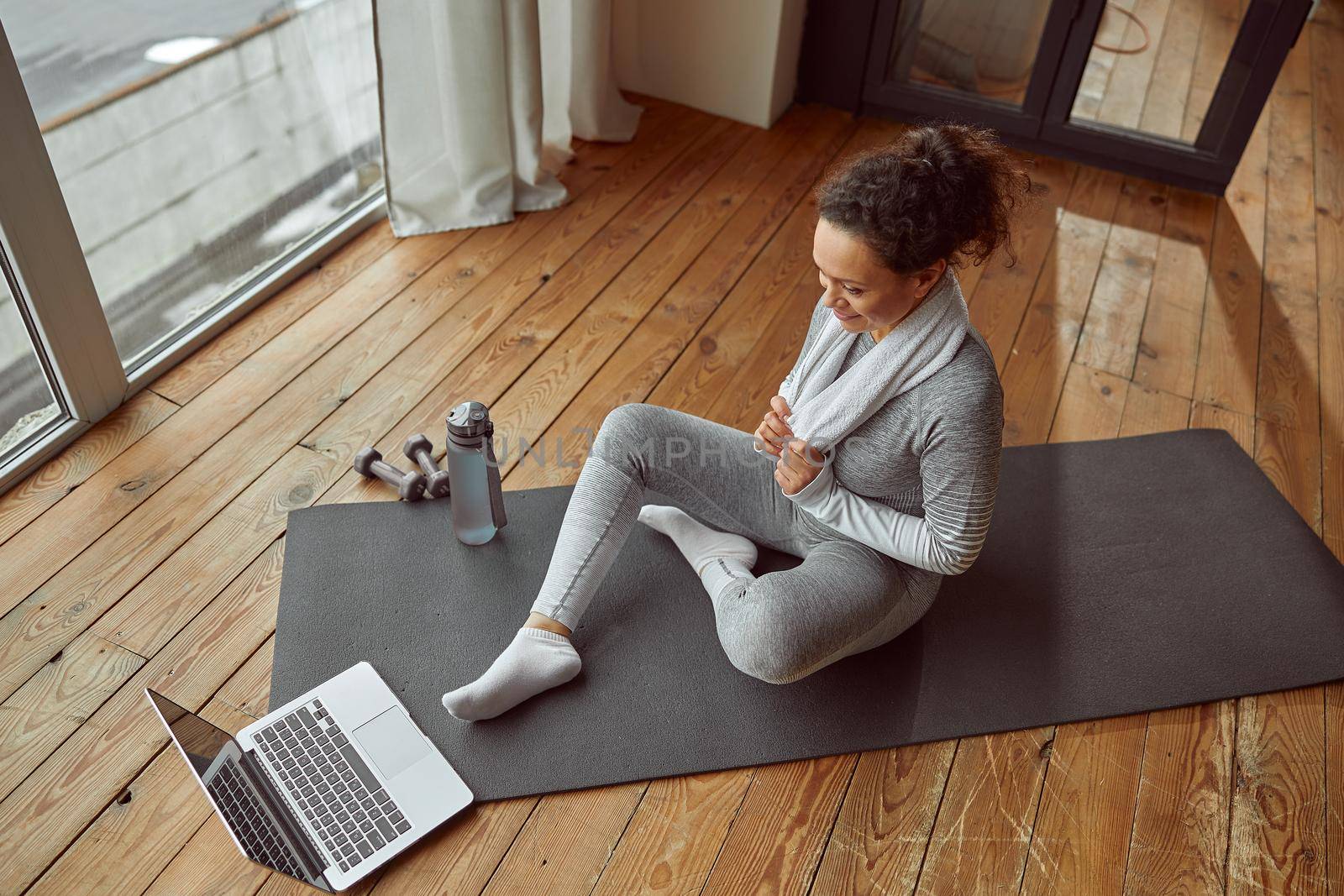 Top view of smiling athletic female exercising on mat while watching video training on laptop