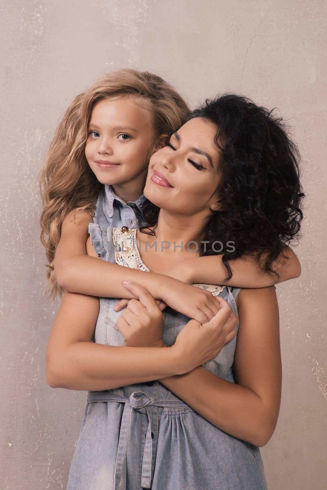 Little girl hugging happy mom from behind near grey wall