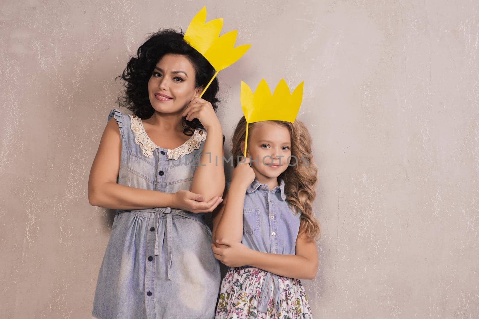 Funny family on a background of gray wall. Mother and her daughter girl with a paper accessories. Mom and child are holding paper crown on stick.