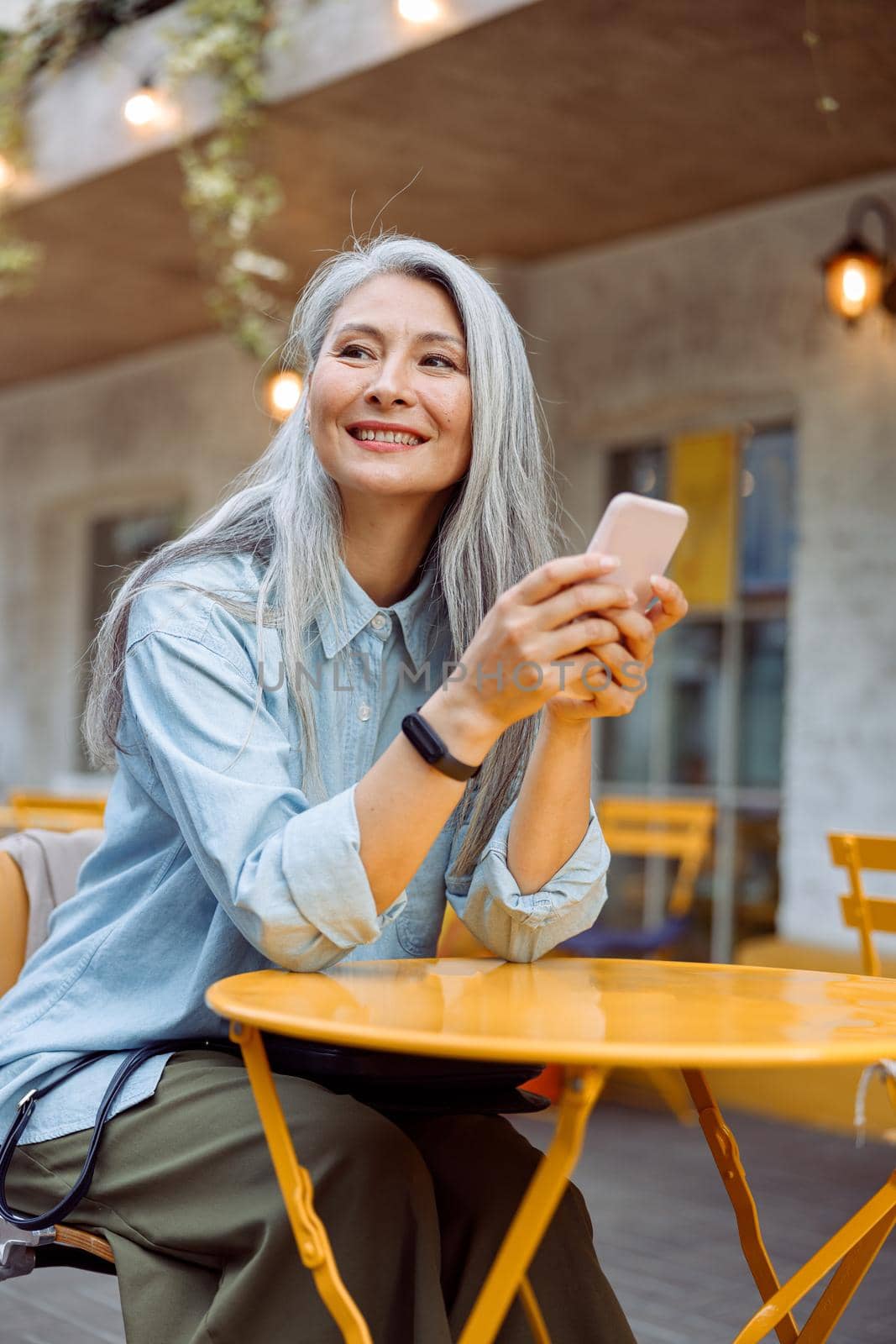 Pretty smiling senior Asian woman holds cellphone at small yellow table on outdoors cafe terrace on autumn day