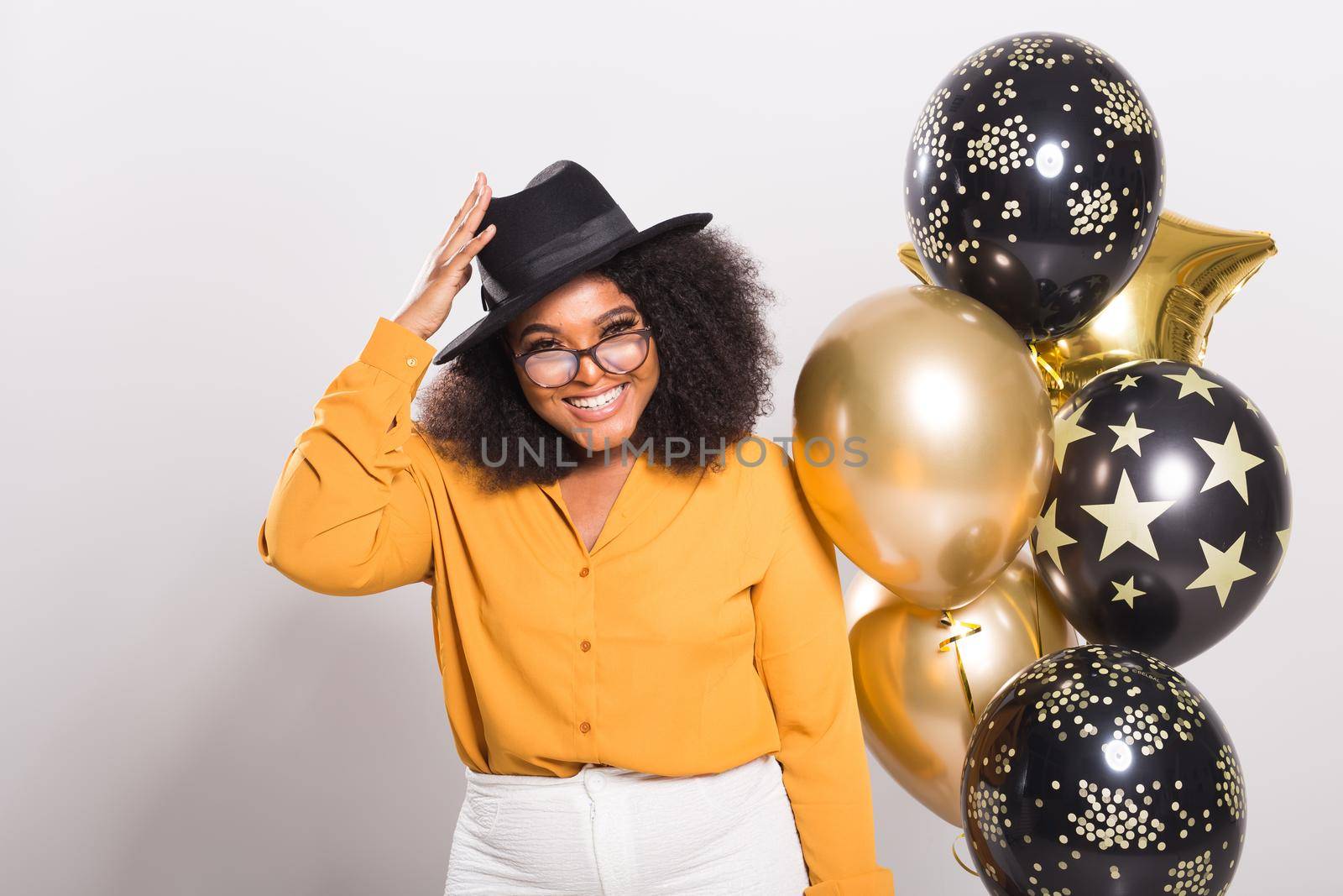 Portrait of smiling young African-American adult woman looking sweet on yellow background holding balloons.