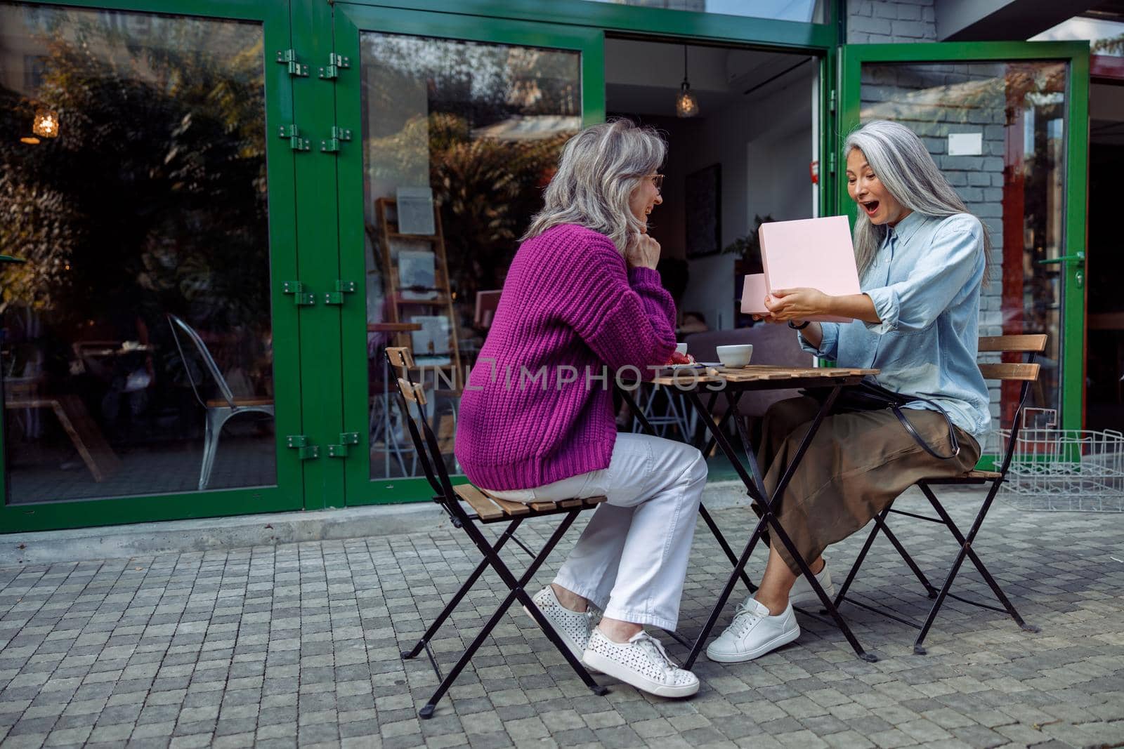 Excited senior Asian woman opens gift presented by best friend sitting at small table on outdoors cafe terrace on autumn day